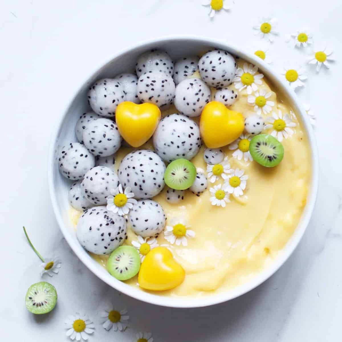 A bowl of yellow smoothie with dragon fruit balls yellow chocolate hearts mini kiwi halves and edible flowers 