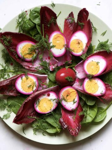 A plate with eight Radicchio leaves and pink halved eggs on top of each leaf with a whole egg in the middle and extra green salad leaves