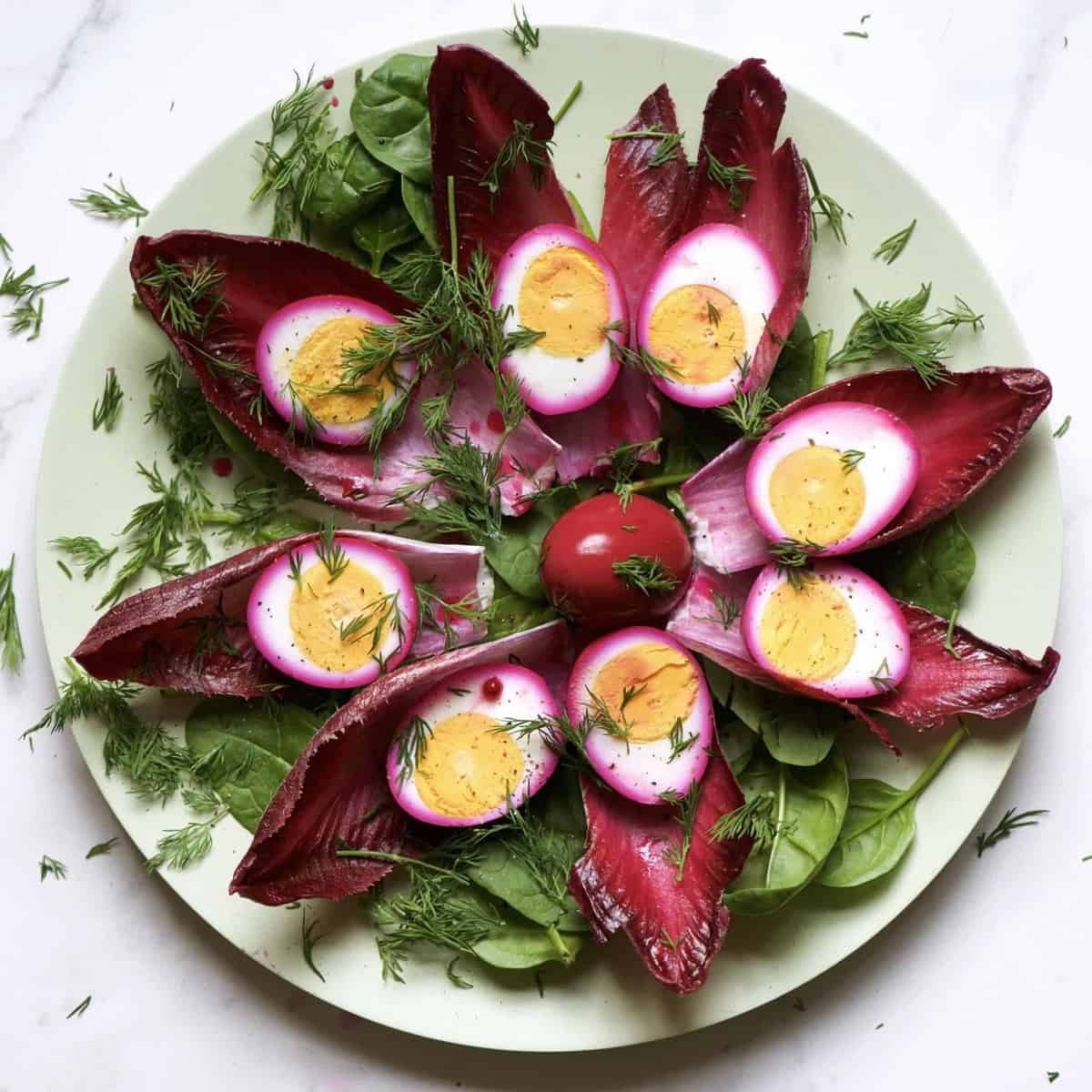 A plate with eight Radicchio leaves and pink halved eggs on top of each leaf with a whole egg in the middle and extra green salad leaves