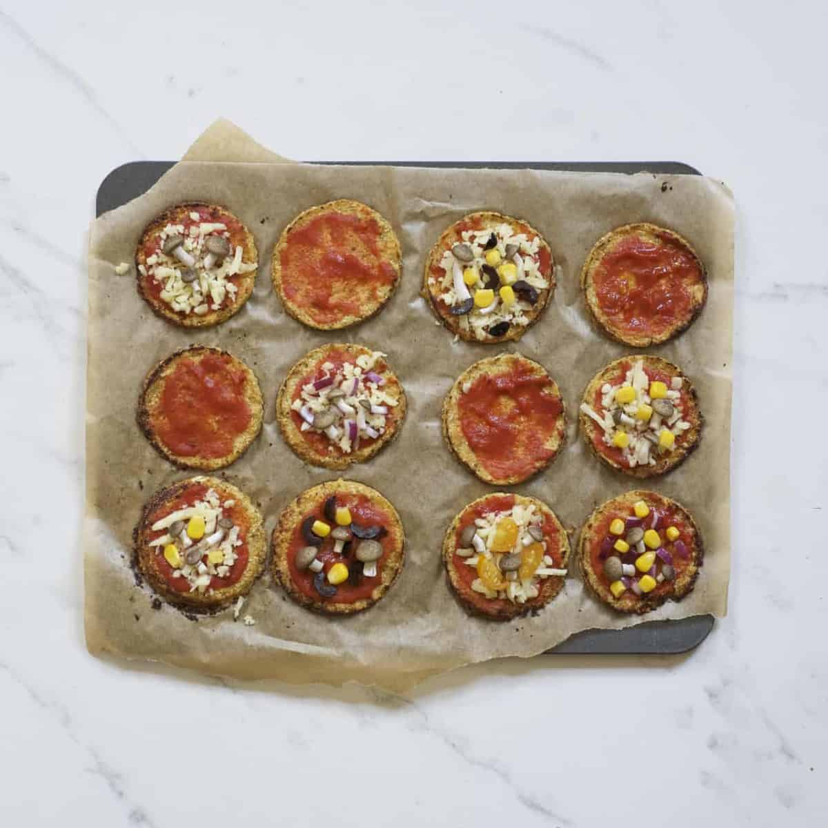 Baked mimi pizzas topped with tomato sauce and some with cheese, corn and mini mushrooms on a baking tray