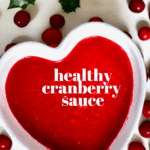 A heart shaped bowl with cranberry sauce and cranberries around it on a flat surface 