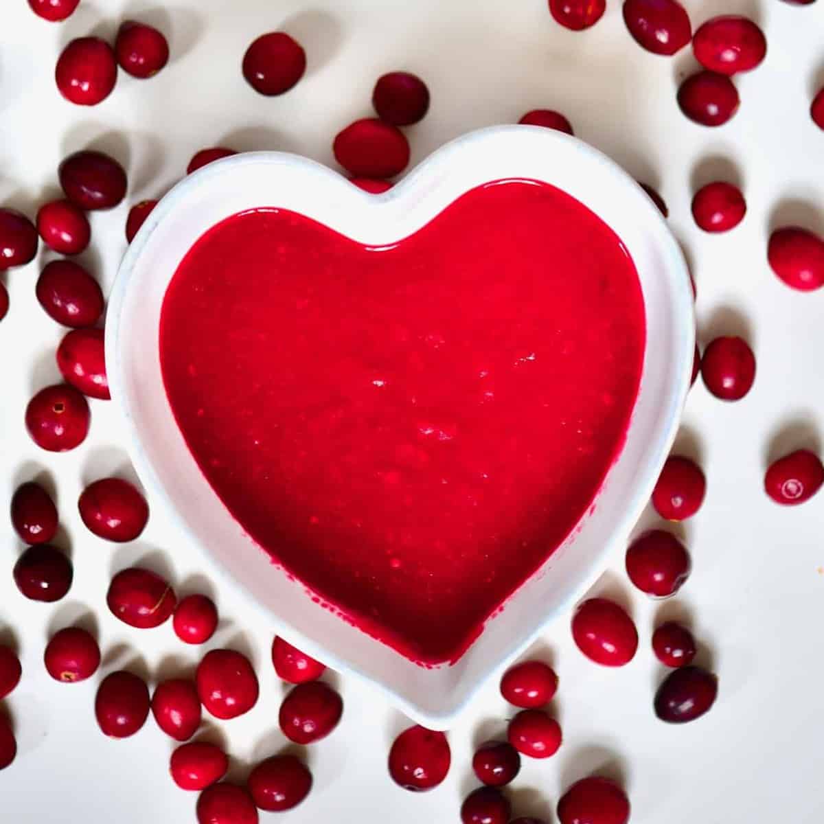 A heart-shaped bowl with cranberry sauce and cranberries around it on a white flat surface 