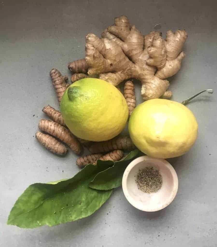 Turmeric and ginger roots with two whole lemons with a leaf and a small bowl with black pepper laying on a flat gray surface