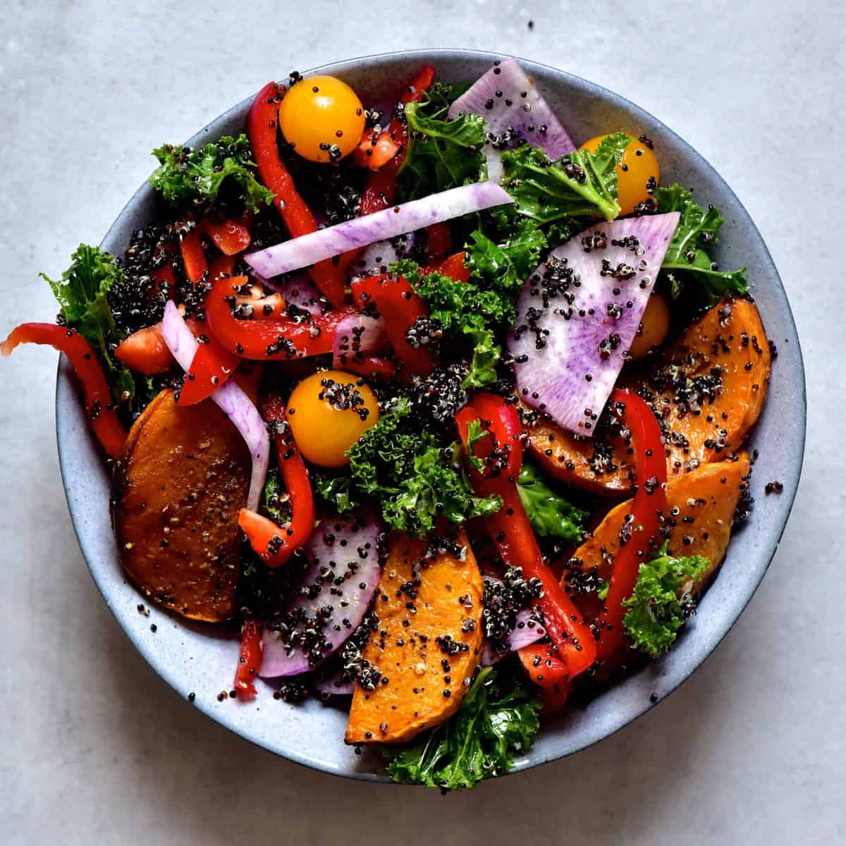 A bowl of butternut squash and kale salad with peppers and tomatoes on a grey background 