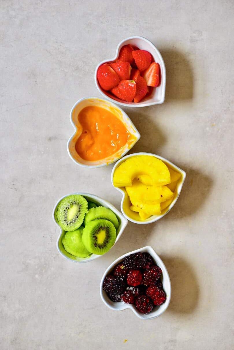 Five bowls filled with strawberries mango pineapple kiwi and blackberries