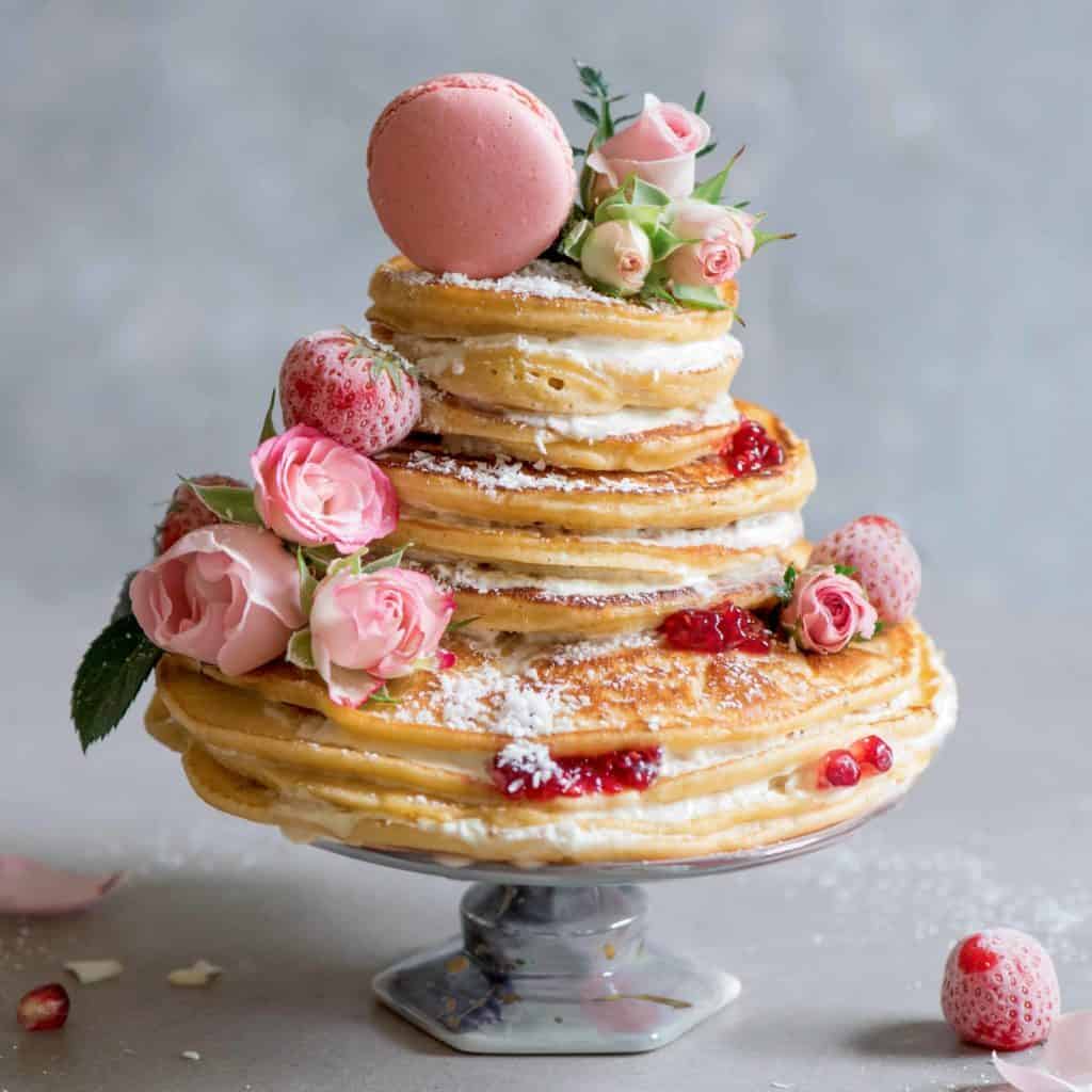 A pancake layered cake decorated with edible roses berries a pink macaroon and shredded coconut