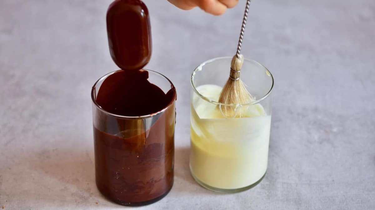 A homemade magnum ice cream over a glass of melted dark chocolate and a glass of white melted chocolate with a little brush