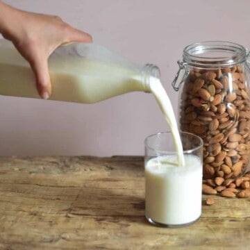 Pouring almond milk from a glass bottle into a glass and a jar or almonds behind the glass