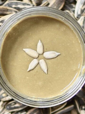 Top view of sunflower seed butter and five sunflower seeds in a jar and sunflower seeds around it on a flat surface