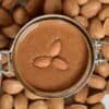 Top view of almond butter and three almonds in a jar and almonds around it on a flat surface
