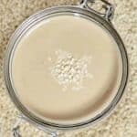 Top view of tahini with some sesame seeds in jar over sesame seeds on a flat surface