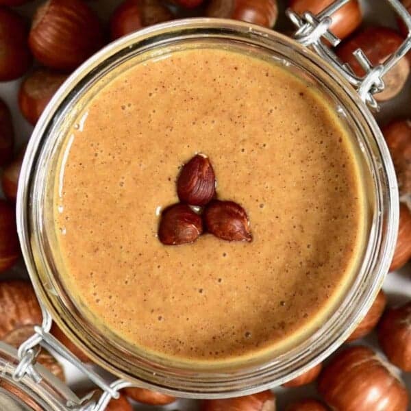 Top view of hazelnut butter and three hazelnuts in a jar and hazelnuts around it on a flat surface