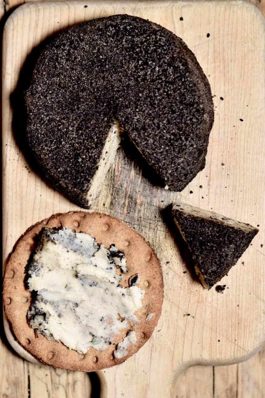 Homemade cashew cheese round with a slice cut off and some spread on a cracker on a wooden board