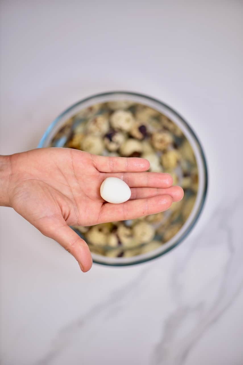 A hand holding one peeled quail egg above a bowl with quail eggs and water