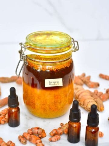 A big jar with homemade turmeric tincture with three small dropper bottles around dit and some turmeric laying on a flat surface