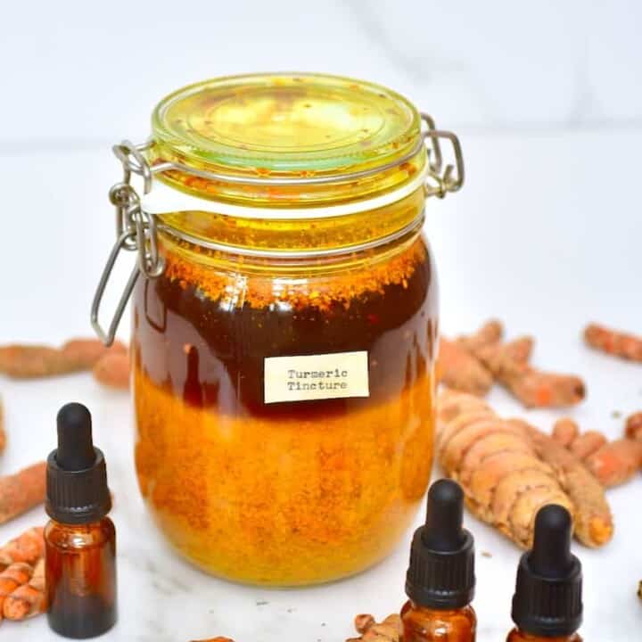 A big jar with homemade turmeric tincture with three small dropper bottles around dit and some turmeric laying on a flat surface
