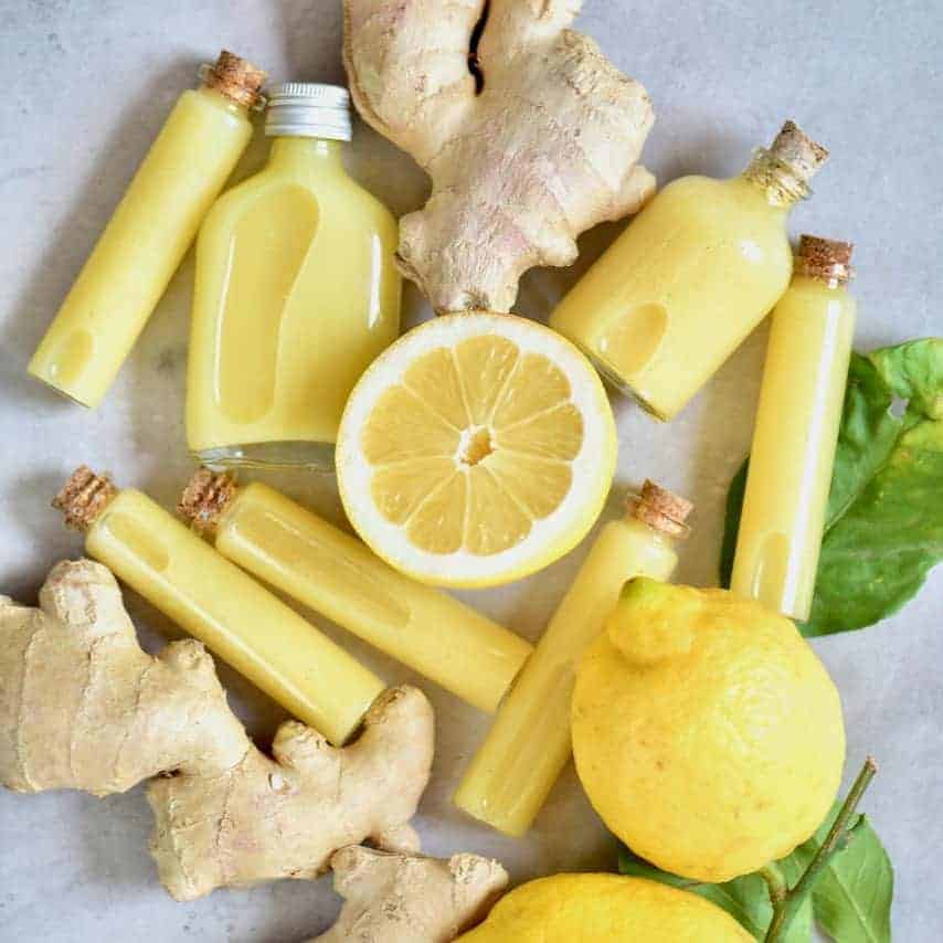 One whole and one half lemon with two ginger roots and 7 small glass vials filled with ginger lemon shots laying on a flat grey surface