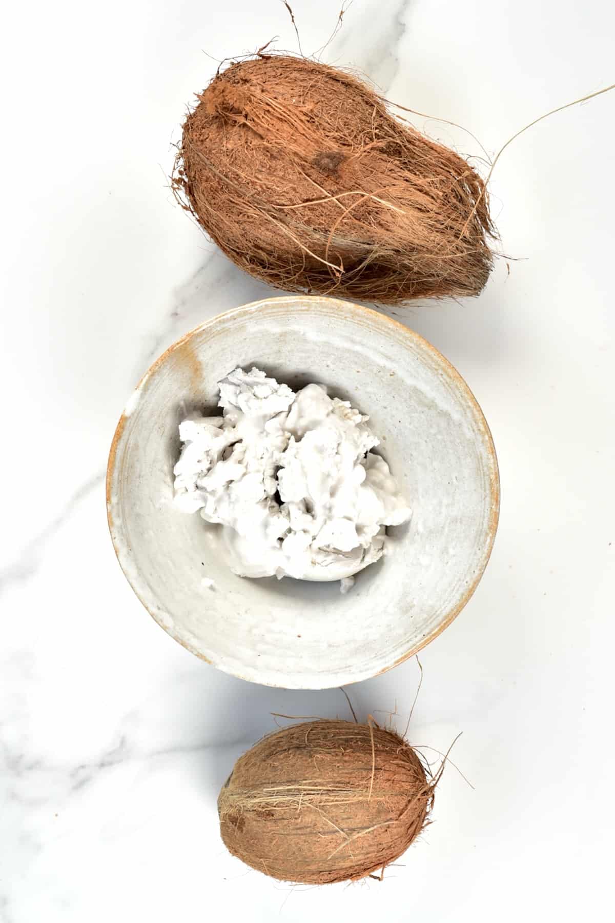 Coconut cream in a bowl and two coconuts on a white surface