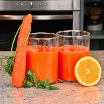 Two glasses of orange carrot juice with a carrot and a slice of orange next to them