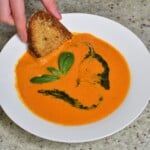 A bowl with tomato soup and a toasted piece of bread being dipping into it