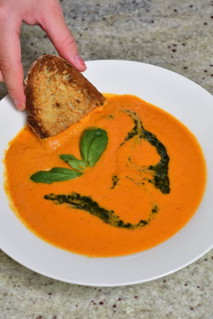 A bowl with tomato soup and a toasted piece of bread being dipping into it