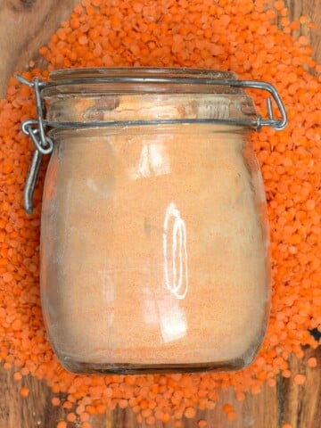 A jar with red lentil flour over some red lentils on a wooden board