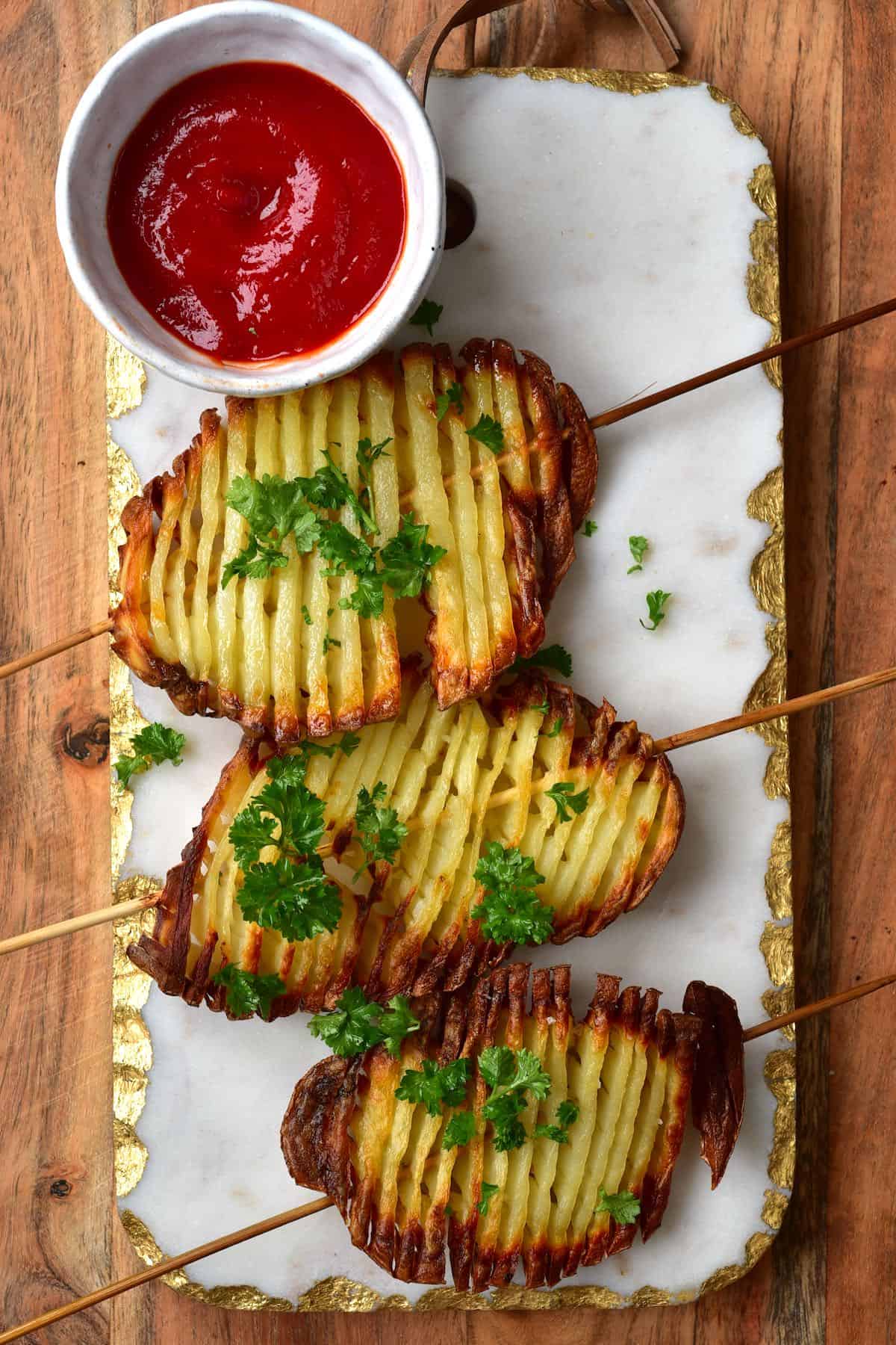 Three accordion potato chips on skewers topped with some parsley and a bowl with ketchup