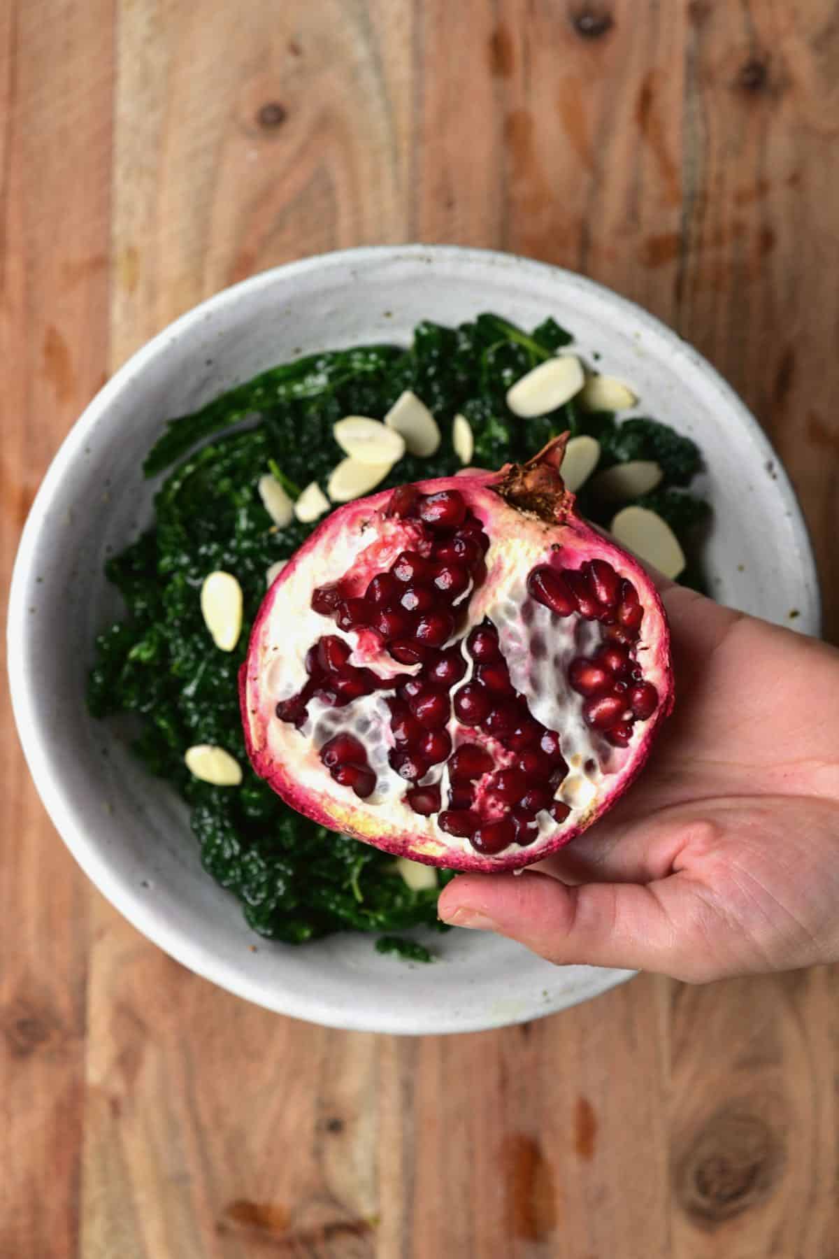 A bowl with kale salad with a half pomegranate being held above it