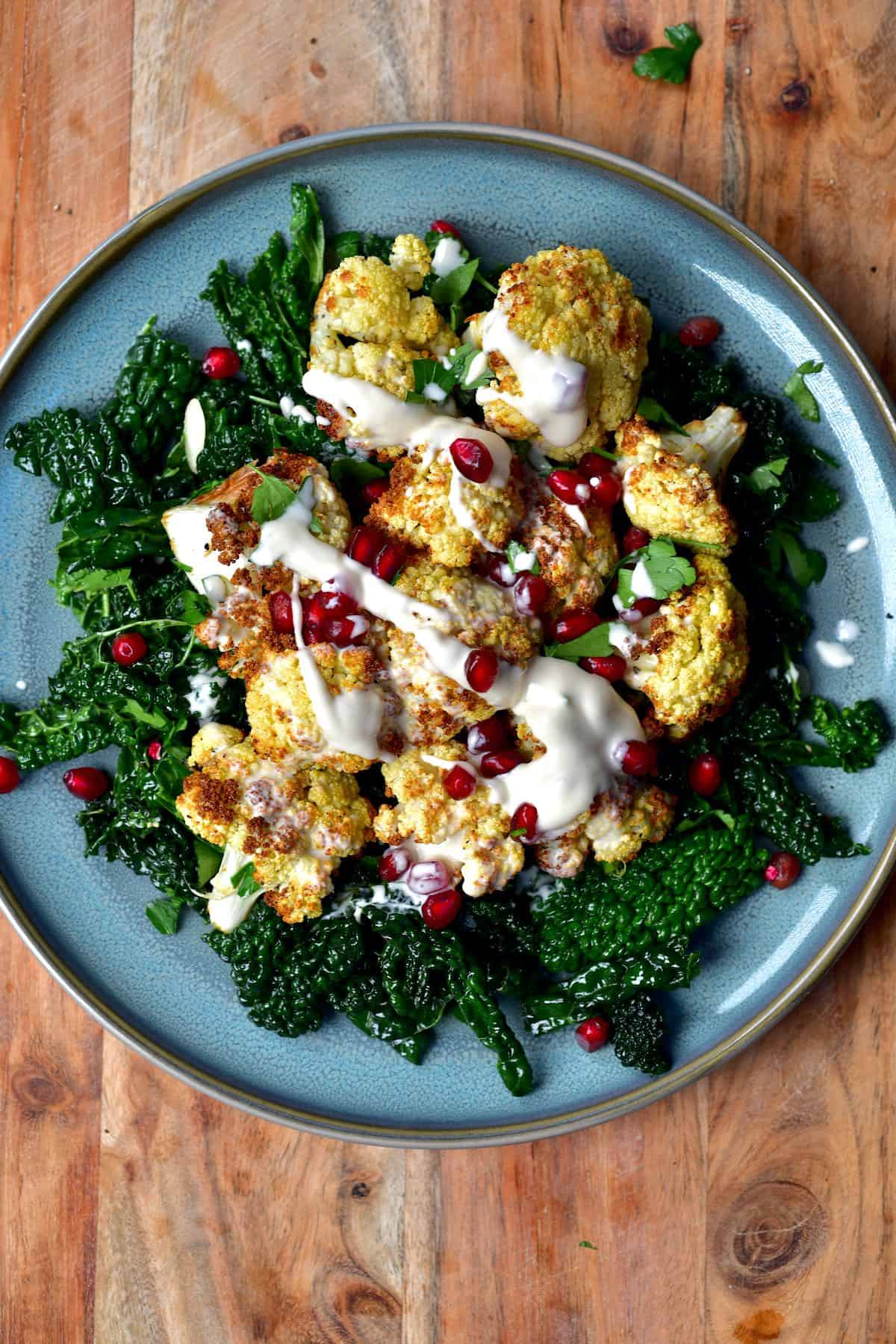 A serving of kale salad and baked cauliflower topped with pomegranate seeds and tahini sauce
