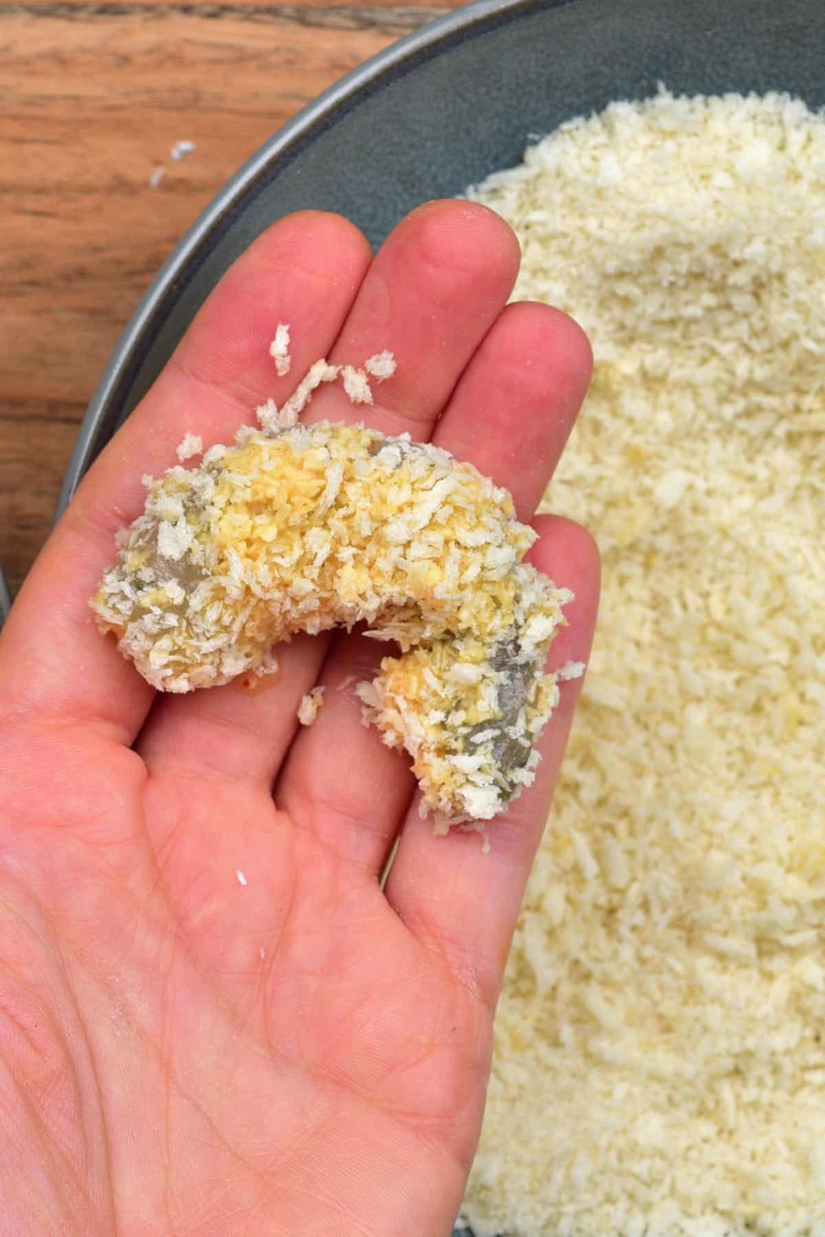 A hand holding a shrimp coated in breadcrumbs and coconut