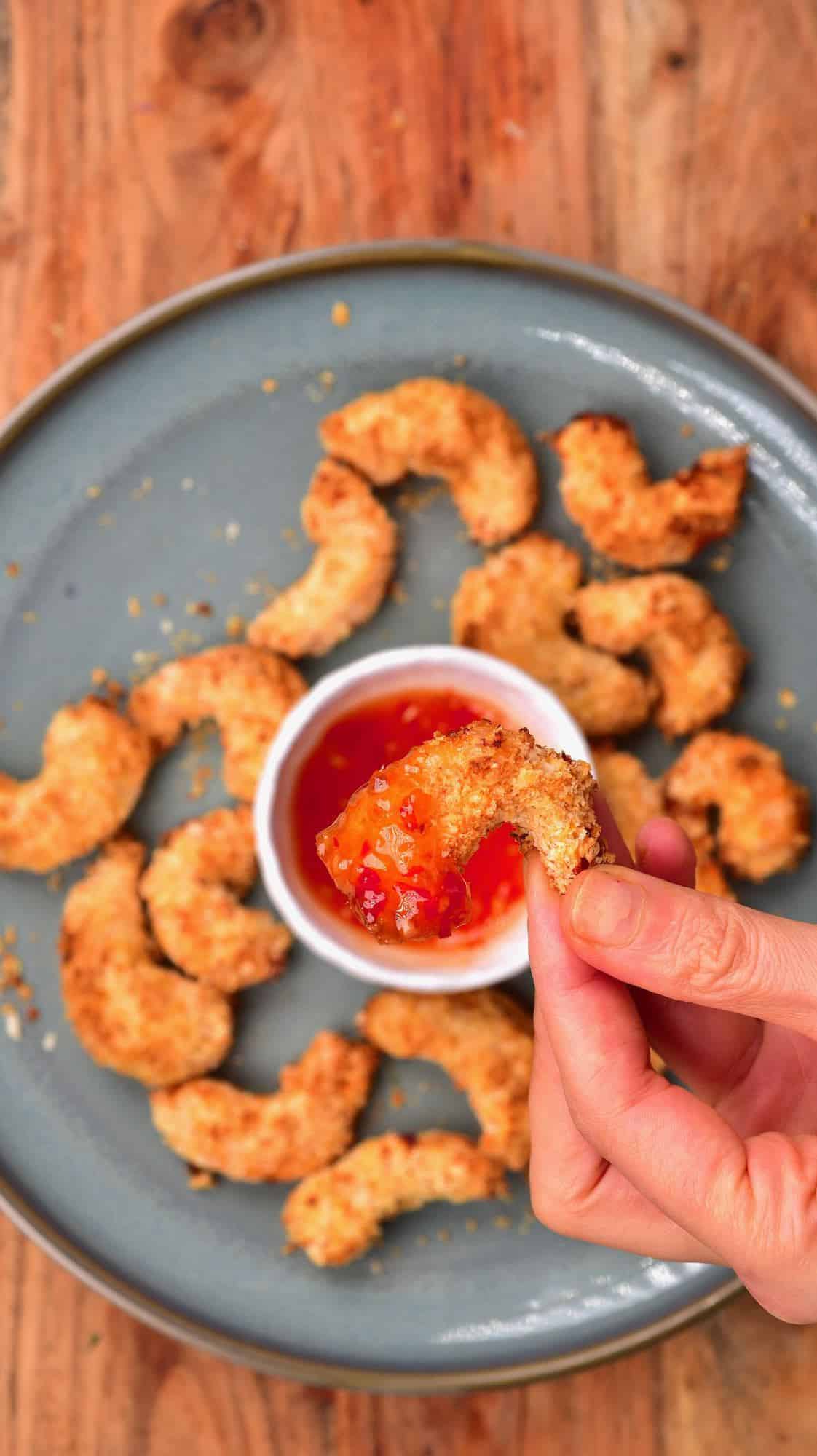 A hand holding a baked shrimp over a bowl with chili sauce in a plate with more baked shrimp