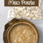 Homemade miso paste in a large container covered with wrap and a bag of beans next to it