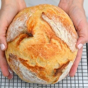 Two hands holding a no-knead bread over a cooling rack