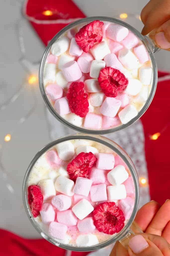 two mugs of white hot chocolate with marshmallows and dried raspberries with lights in the background