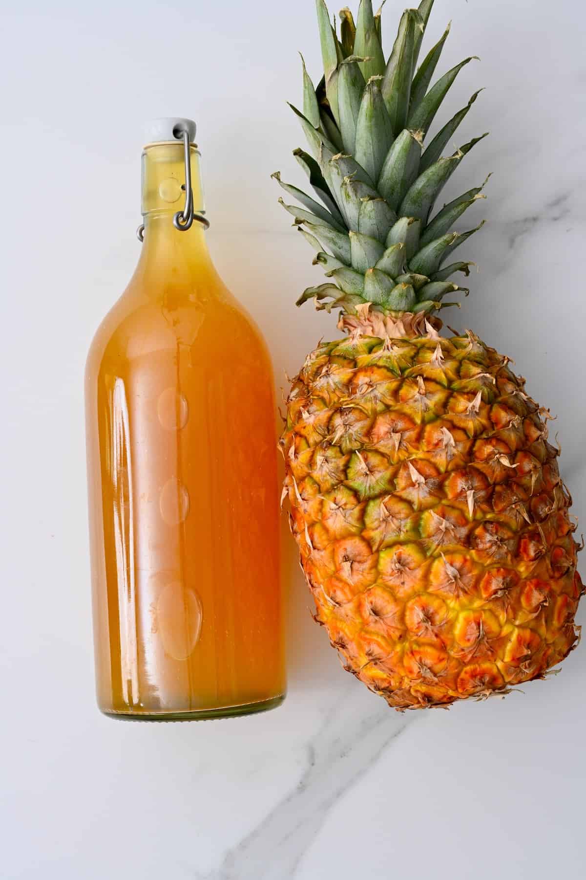A bottle of tepache next to a pineapple laying on a flat surface