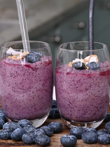 Two glasses with blueberry smoothie with straws and blueberries around them