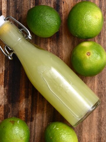A small bottle with lime juice and limes on a chopping board