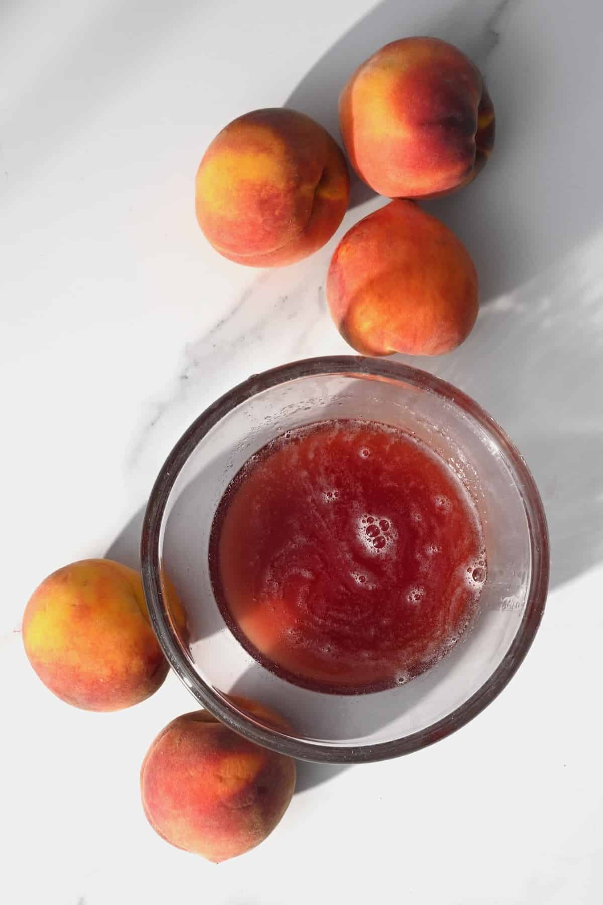 A bowl with peach syrup and five peaches next to it