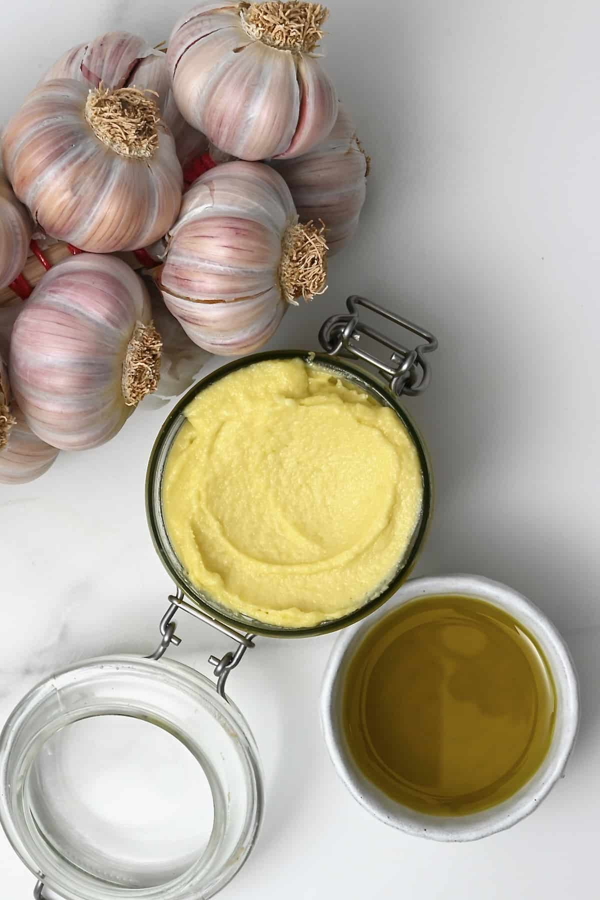 Homemade garlic aioli in a jar next to garlic heads and olive oil