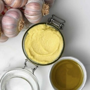 Homemade garlic aioli in a jar next to garlic heads and olive oil