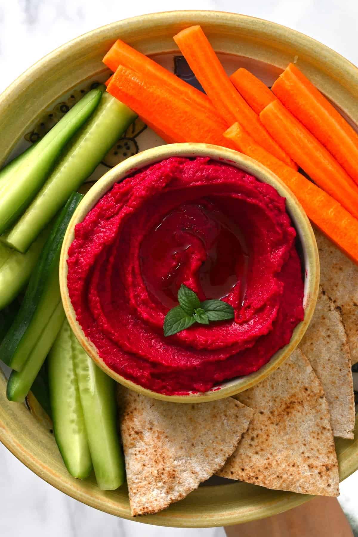 Beet hummus in a small bowl with cucumber and carrot sticks and pita chips