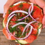 Hand holding a Lox bagel topped with dill capers and onion