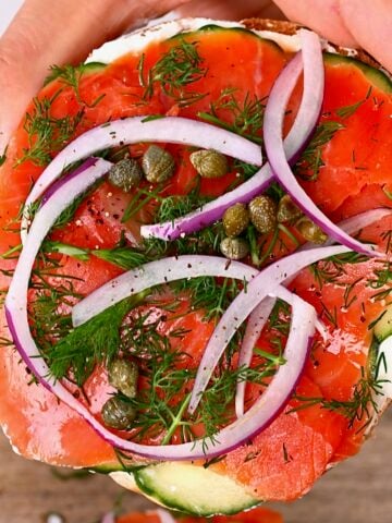 Hand holding a Lox bagel topped with dill capers and onion