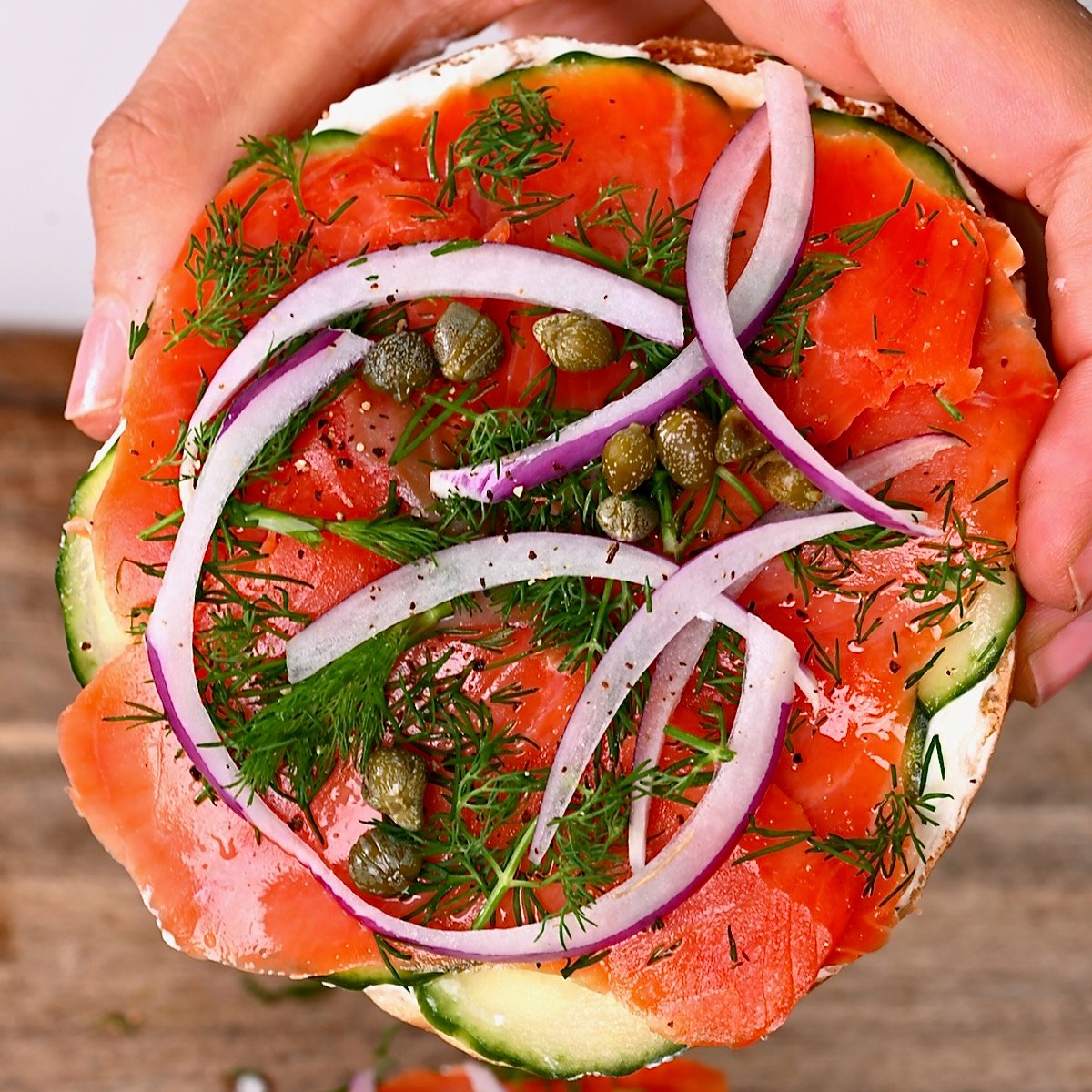 Hand holding a Lox bagel topped with dill capers and onion