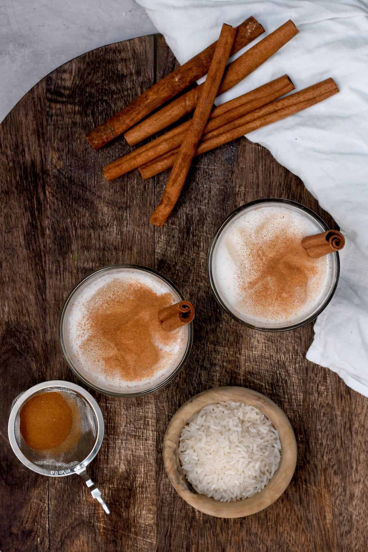 Top view of two glasses with horchata and a small bowl with rice, few cinnamon sticks and cinnamon powder