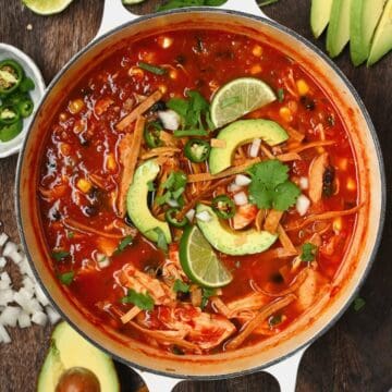 Homemade chicken tortilla soup in a large saucepan topped with avocado and lime wedges