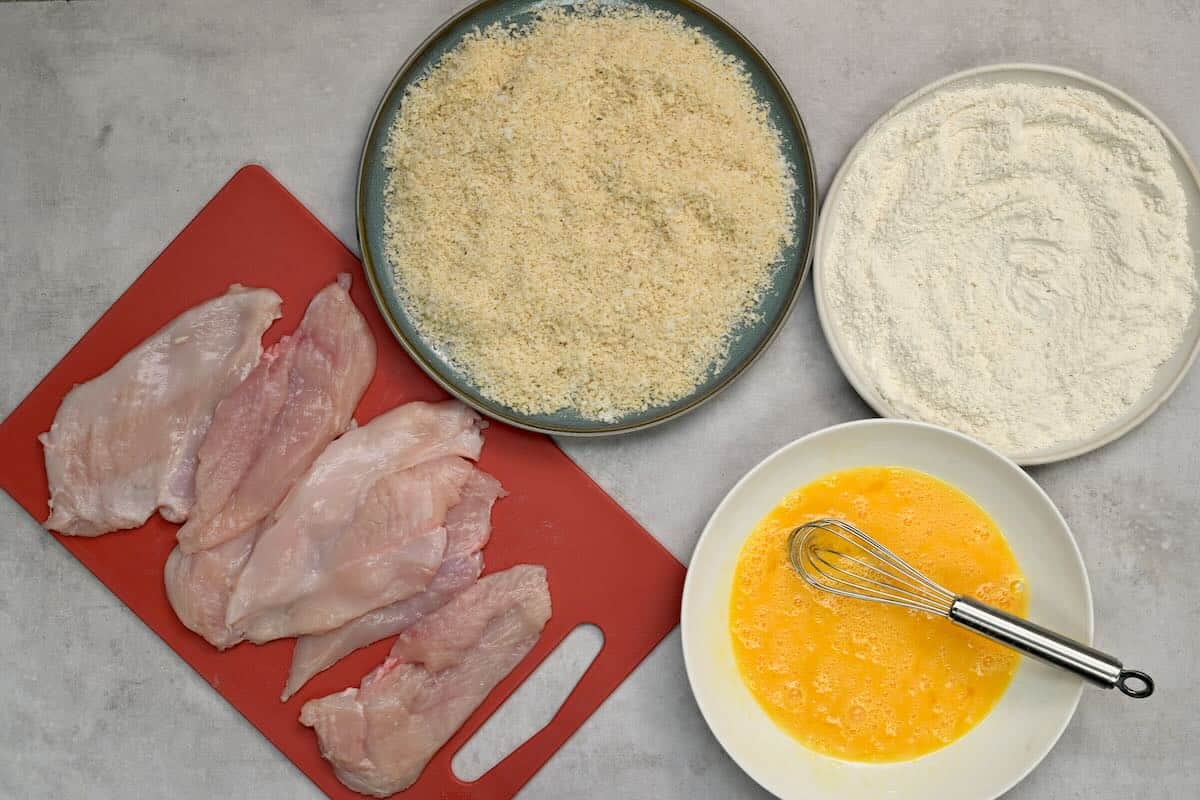 Arranging the coating station with flour, breadcrumb, and egg mixture alongside the prepared chicken
