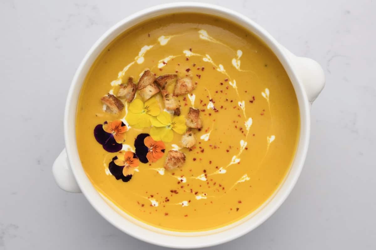 A serving bowl with carrot cream soup topped with croutons and edible flowers