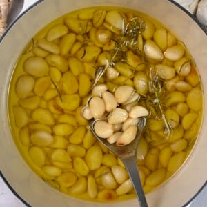 A saucepan with homemade garlic confit and a spoon removing some cloves and oil.