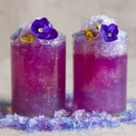 Butterfly pea color changing lemonade served in two glasses with lots of ice, topped with fresh edible flowers.